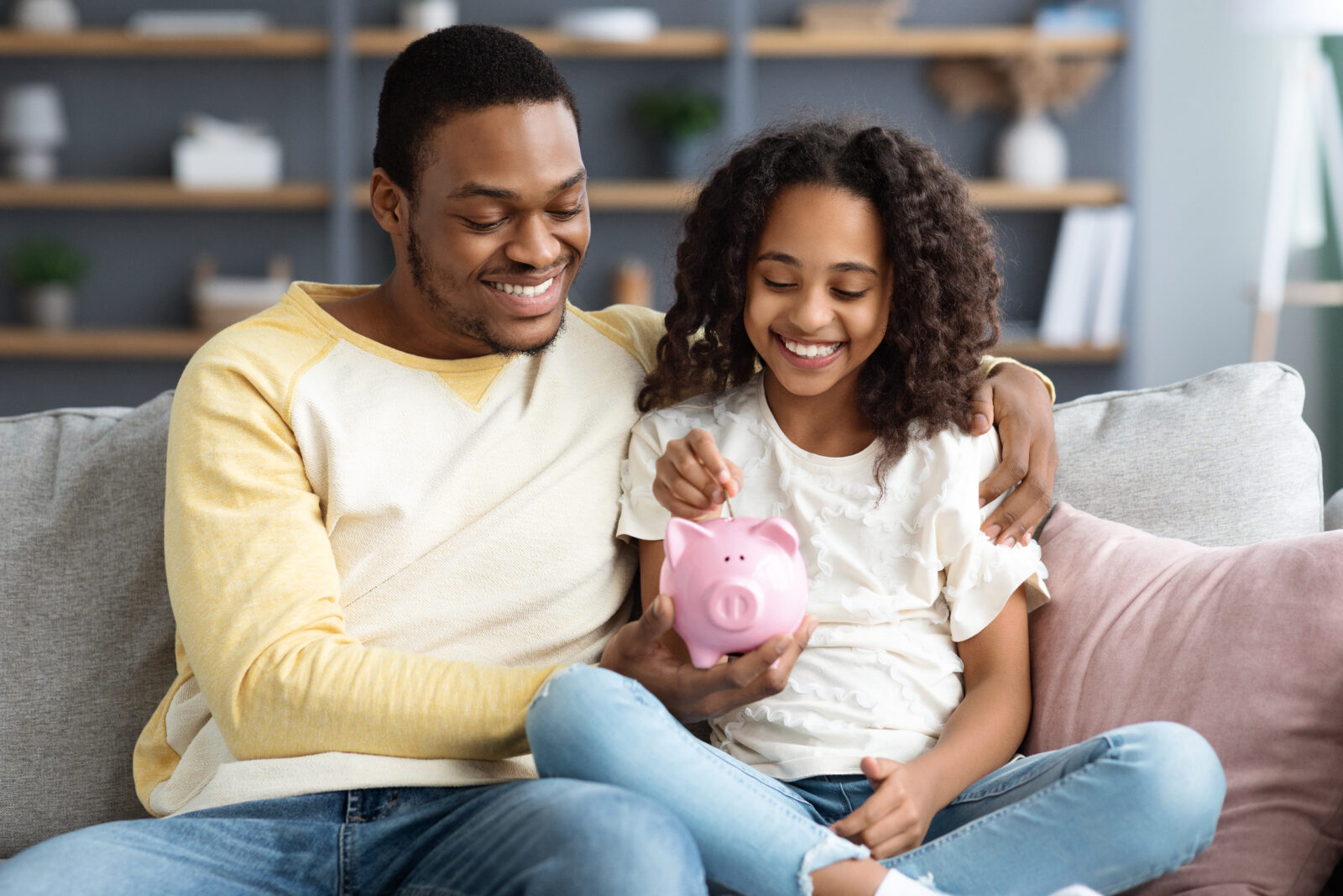 Black girl and father putting coin into piggy bank, sitting on sofa at home. Happy african american dad teaching his cute daughter how to save money, living room interior. Financial education concept
